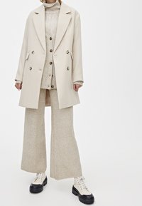 Cappotto di lana beige doppiopetto indossato sopra un maglione in maglia beige e pantaloni a gamba larga coordinati, abbinati a stivali piattaforma bianchi e neri.