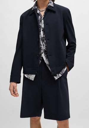 Person trägt eine schwarze, geknöpfte Jacke über einem schwarz-weiß gemusterten Hemd, passende schwarze Shorts und eine silberne Kettenhalskette.