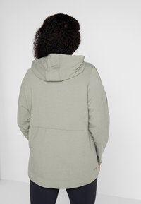 Sweat à capuche vert clair avec une coupe décontractée, présentant une texture de tissu et un design simple au dos, s'étendant jusqu'aux hanches.
