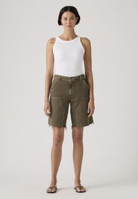 Γυναίκα που στέκεται, φοράει λευκό τοπ χωρίς μανίκια, λαδί βερμούδα μέχρι το γόνατο με μεγάλες μπροστινές τσέπες και μαύρα σαγιονάρες σε ουδέτερο φόντο.