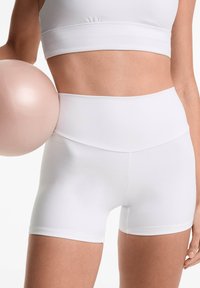 White athletic shorts with a high waist, made of smooth fabric. Paired with a matching crop top, holding a light pink exercise ball.