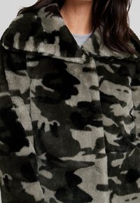 Abrigo de piel sintética con un patrón de camuflaje, con un cuello ancho, textura suave y una combinación de tonos negros y grises.