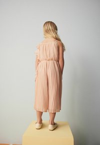 Robe plissée pêche avec épaulettes volantées, taille cintrée et jupe midi. Portée avec des chaussures légères à enfiler.