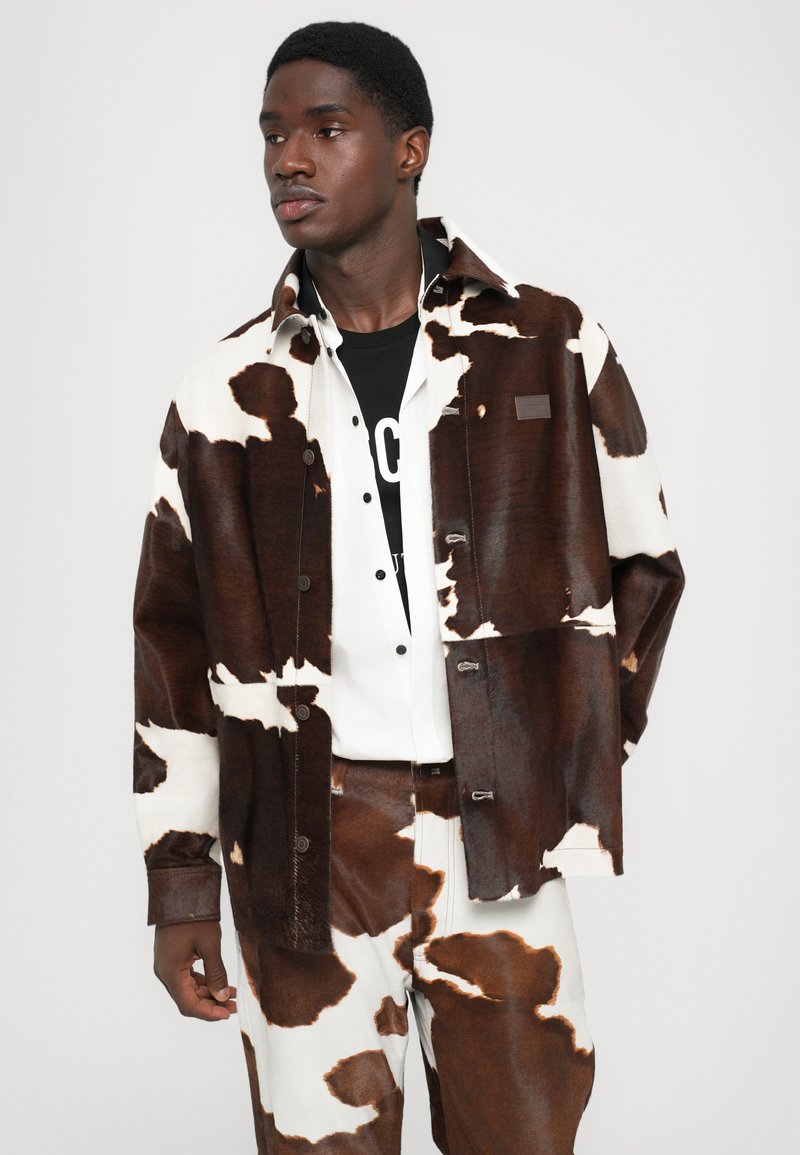 Braune und weiße Kuhfleckenjacke mit passenden Hosen, aus strukturiertem Material gefertigt. Verfügt über Druckknöpfe und ein dezent gesetzt Logo-Patch.
