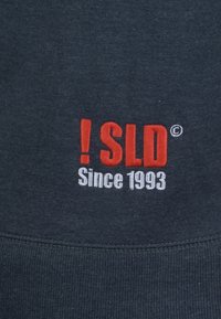 Tmavě modrá tkanina mikiny s výrazným červeným a bílým vyšitým textem: „!SLD“ nad nápisem „Since 1993“. Text je umístěn uprostřed na prsou.