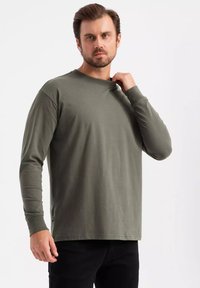 T-shirt a maniche lunghe color verde oliva in cotone. Presenta un collo a girocollo e una leggera elasticità con una vestibilità rilassata. Design minimalista, senza motivi.