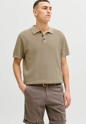 Bărbat tânăr purtând un tricou polo bej din tricot cu mâneci scurte și pantaloni scurți maro, stând cu o mână în buzunar, uitându-se în lateral pe un fundal simplu.