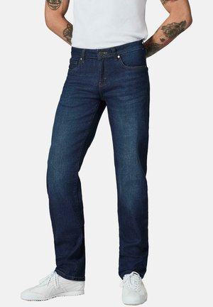 Mann trägt dunkelblaue Jeans mit geradem Bein, weiße Sneakers und ein weißes ärmelloses Shirt, zeigt Tätowierungen auf beiden Armen.