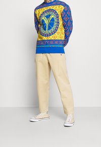 Felpa con motivo blu e giallo, dotata di design grafico, abbinata a pantaloni beige e sneaker chiare. Caratterizzata da colori vivaci e dettagli intricati.