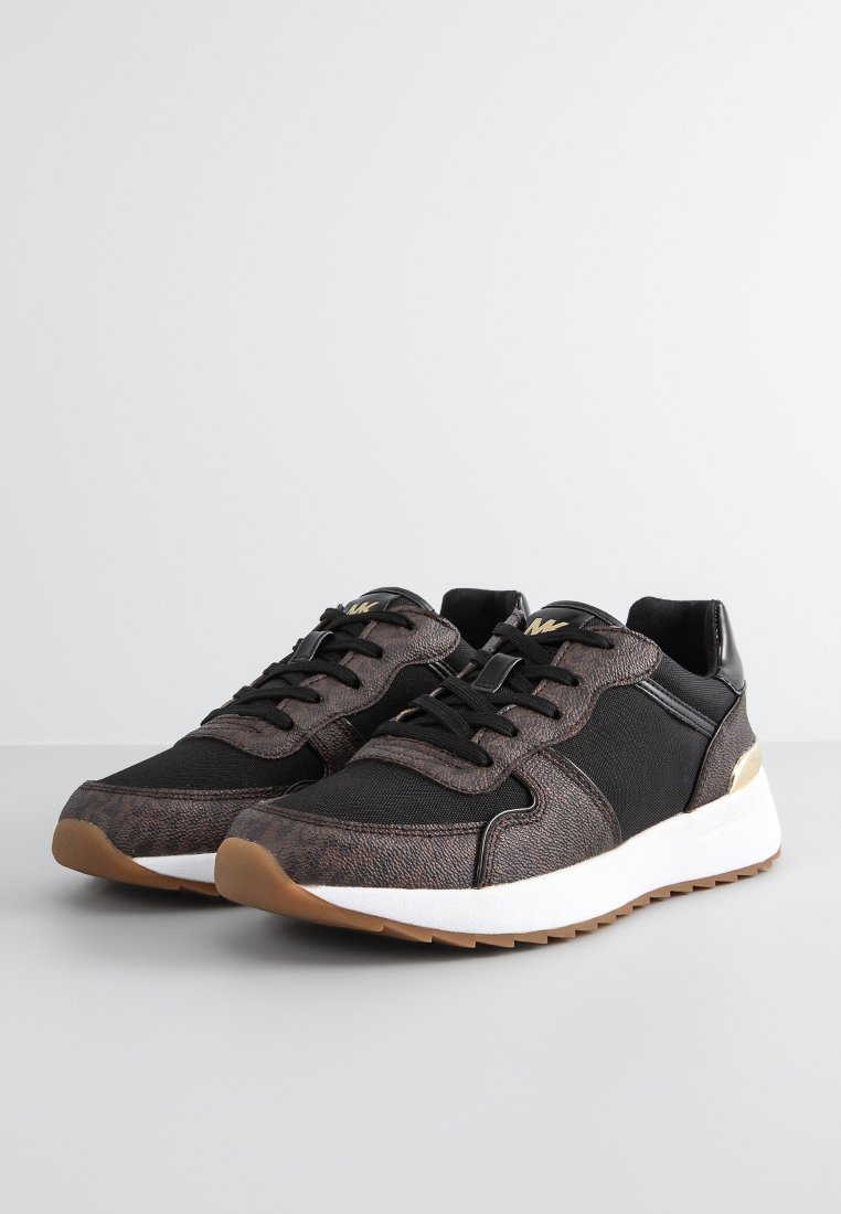 Une paire de baskets texturées noires et marron foncé avec des semelles blanches et une semelle en couleur gomme, exposée sur une surface gris clair.