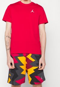 Hombre vistiendo una camiseta roja de Jordan y pantalones cortos de patrón zigzagueante multicolor, de pie contra un fondo blanco.