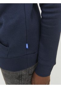 Sweatshirt bleu marine avec poignets et bas côtelés, présentant une petite étiquette bleue sur le côté. Texture douce, design ajusté et longueur standard.