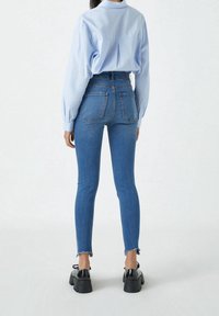 Jeans ajustados en denim azul medio con cintura alta, con bolsillos traseros y dobladillo sin terminar. Combinados con una camisa de botones azul claro.