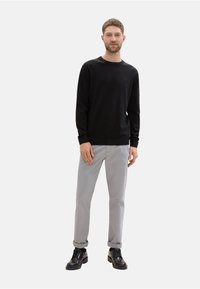 Schwarzer langärmeliger Pullover, hellgraue Hose, schwarze Schuhe. Das Model steht aufrecht, mit minimalem Hintergrund, die Arme entspannt an den Seiten.