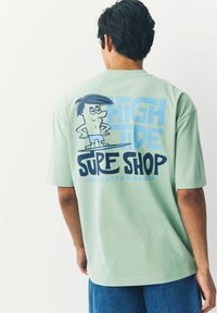 Mint grøn oversized t-shirt med et tegneseriefigur grafik på ryggen, med blå tekst der står "HIGH TIDE SURF SHOP MANHATTAN BEACH."