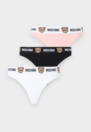 Žena nosiaci biely bralet Moschino a zodpovedajúce nohavičky s pásmi s logom medvedíka, stojaca s rukami za chrbtom.