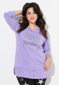 Tunic a maniche lunghe lilla con orlo arrotondato. Presenta una scollatura con bottoni e strass argentati che formano la scritta "RISE" con un design che gocciola.