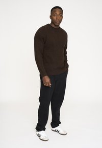 Brun texturerad tröja, svarta byxor och vita sneakers med svarta ränder. Modellen står vänd mot kameran med en neutral uttryck mot en vit bakgrund.