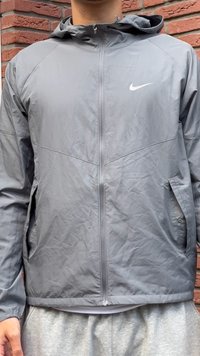 Persona che indossa una giacca a zip con cappuccio grigia della Nike e pantaloni sportivi grigi chiari, in piedi di fronte a un muro di mattoni.