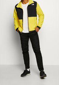 Černá a žlutá fleecová bunda se zipem na přední straně, s dvěma kapsami na zip, kombinovaná s černými kalhotami typu cargo a černými teniskami.