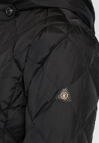 Veste matelassée noire avec un design matelassé, une texture brillante et un écusson logo triangulaire sur la manche. Fermeture à boutons au niveau du col.