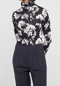 Black turtleneck top featuring floral print in white and light gray, paired with high-waisted dark gray trousers. Smooth fabric, fitted design.
