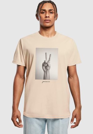 T-shirt en coton beige avec un graphique noir et blanc d'une main faisant un signe de paix, avec le mot "paix" dans une police élégante en dessous.