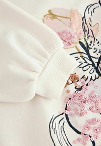 Hanorac alb din bumbac, cu broderie florală în nuanțe de roz și marron, finisaj texturat și paiete roz împrăștiate pe material.