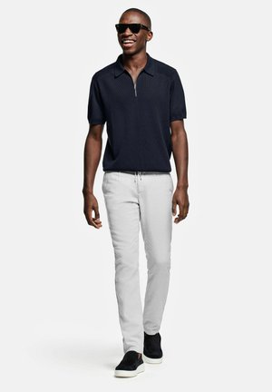 Homme souriant portant des lunettes de soleil noires, une chemise bleu marine à manches courtes avec col zippé, un pantalon gris clair et des chaussures noires sans lacets marchant vers l'avant.