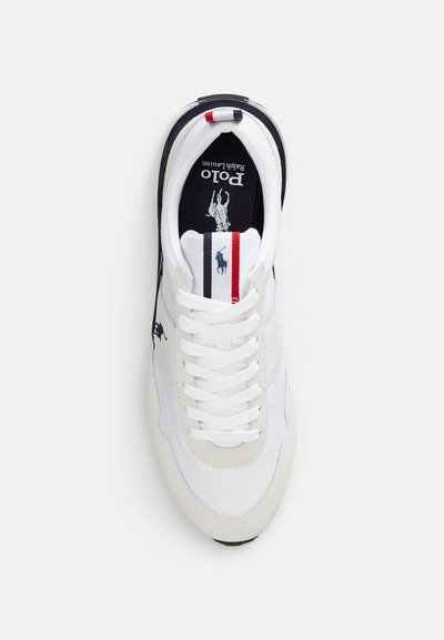 Fehér sportcipő sima textil és velúr anyagok keverékével, fekete logóval, piros és kék részletekkel a saroknál, valamint fehér fűzőkkel.