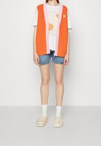 Henkilö, jolla on päällään oranssi neulottu liivi pinkillä reunuksella, valkoinen kukkakuviollinen t-paita, farkkushortsit, valkoiset sukat ja beigekat slaidikengät.