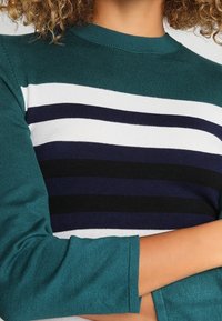 Stickad tröja med en teal-färgad kropp och 3/4 ärmar, med horisontella ränder i vitt, marinblått och svart över bröstet.