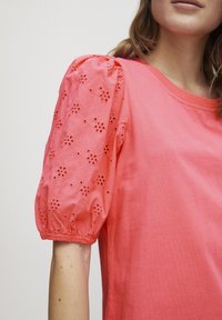 Top din bumbac coral cu mâneci bufante, decorat cu broderie florală și manșete elastice. Guler rotund și croi lejer. Textură netedă și moale.