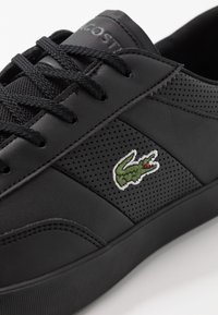 Baskets en cuir noir avec panneau latéral perforé, lacets noirs et logo crocodile vert brodé sur le côté.