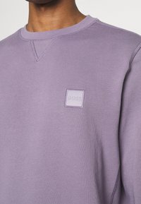 Homme portant un sweat lavande à col rond avec un petit patch logo carré "BOSS" sur la poitrine gauche.