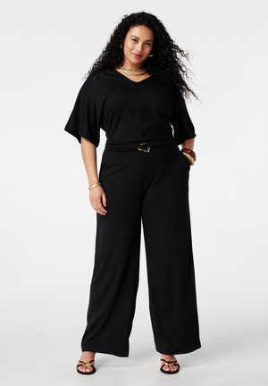 Vrouw met lang krullend haar, gekleed in een zwarte jumpsuit met wijde pijpen en gouden sieraden, staand met één hand in haar zak tegen een witte achtergrond.