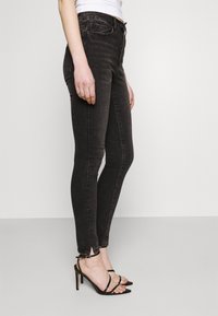 Mujer vistiendo jeans ajustados negros con aberturas en los tobillos y sandalias de tacón alto negras con tiras, de pie contra un fondo liso.