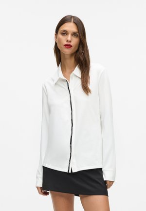 Cămașă albă cu mânecă lungă, având un sistem de nasturi negru, asortată cu o fustă mini neagră. Textură netedă, design clasic al gulerului, stil minimalist.