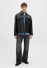 Sort læderjakke med store lommer, parret med en blå denimskjorte og brede mørke jeans. Modellen står foran en ensfarvet baggrund.