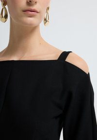 Musta olkaimeton mekko suoralla pääntiellä, jossa on kapeat hihnat. Kullansävyiset pisaramuotoiset korvakorut tuovat metallista korostusta. Silkkinen rakenne.