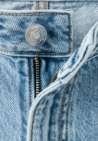 Gros plan sur la fermeture éclair partiellement ouverte d'un jean en denim bleu avec un bouton en métal portant l'inscription « Authentic MNG Denim ».