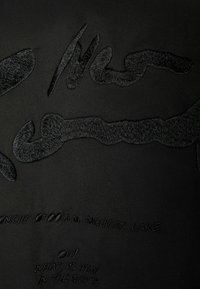 Svart tyg med upphöjd texturerad broderi som har abstrakta mönster och text, vilket skapar en lager-på-lager visuell effekt.