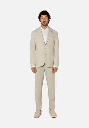 Mann steht, trägt einen hellbeigen Anzug mit weißem Hemd und beigen Slip-on-Schuhen vor einem einfarbig hellen Hintergrund.