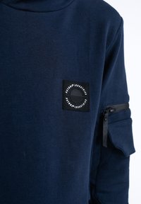 Sweat-shirt bleu marine en coton avec un col montant, doté d'un patch carré noir et d'une poche zippée sur la manche.