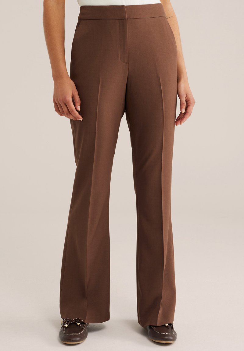 Pantalon marron taille haute, ajusté, avec des plis à l'avant, porté avec des mocassins marron foncé sur un fond neutre.