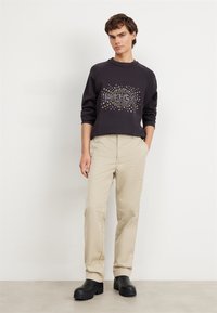 Sudadera negra con gráfico metálico "HUGO", pantalones beiges y zapatos negros gruesos. La tela es suave; el diseño cuenta con un cuello acanalado.