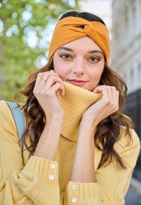 Maglione giallo a maglia con colletto alto a coste e una fascia torcigliata color senape. Texture liscia, bottoni ai polsini, palette di colori calda.