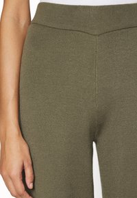 Leggings de malha verde oliva com um largo cós elástico e textura sem costura, apresentando costura vertical ao longo do lado.