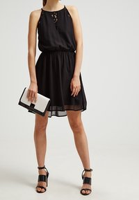 Robe noire sans manches avec encolure en dentelle et taille froncée. Accompagnée d'une pochette blanche et noire et de sandales à talons noires.