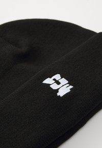 Black knit beanie featuring a textured fabric. The front displays a prominent white embroidered logo in a stylized font.